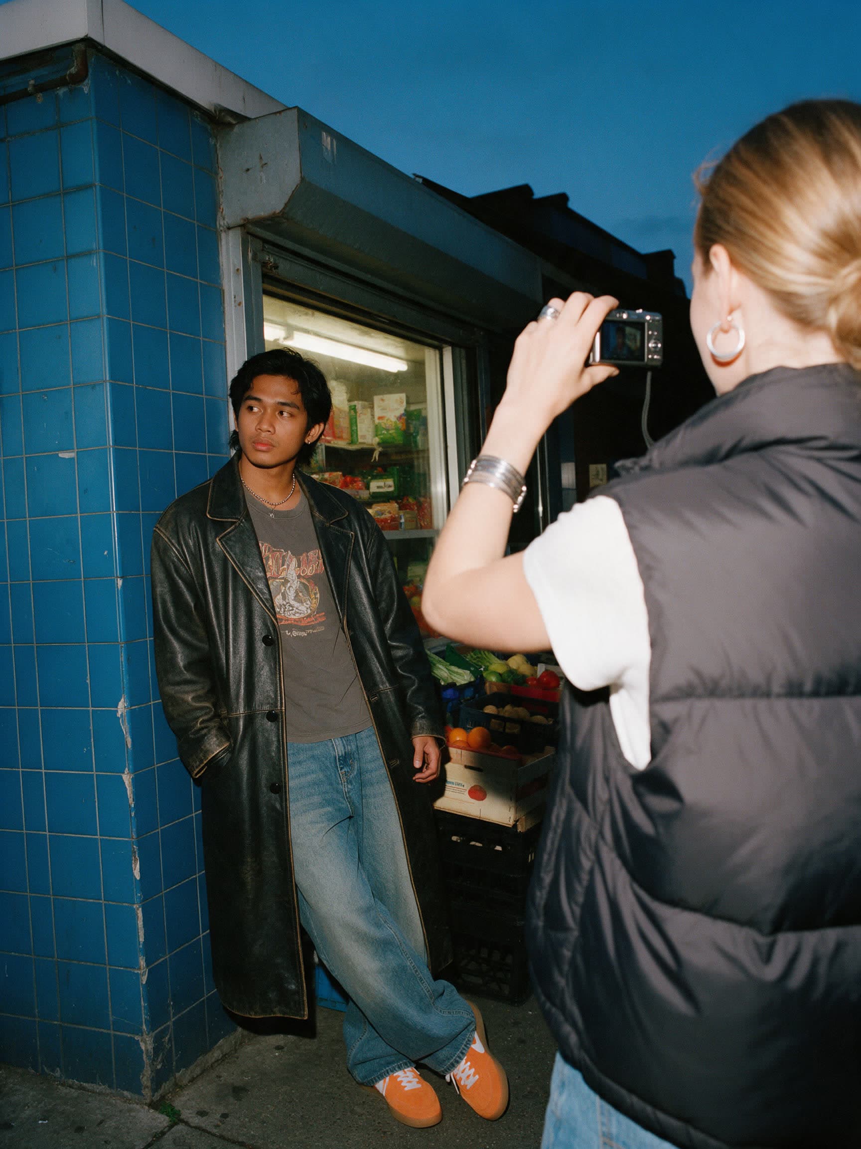 A creator photographing a friend outside a corner shop at dusk