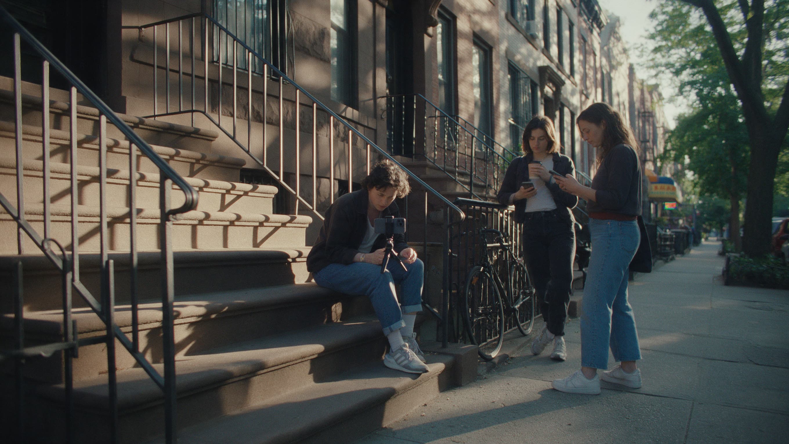Three friends at a brownstone stoop in late light: one filming from the steps on a phone tripod, two on the sidewalk with a coffee and a phone